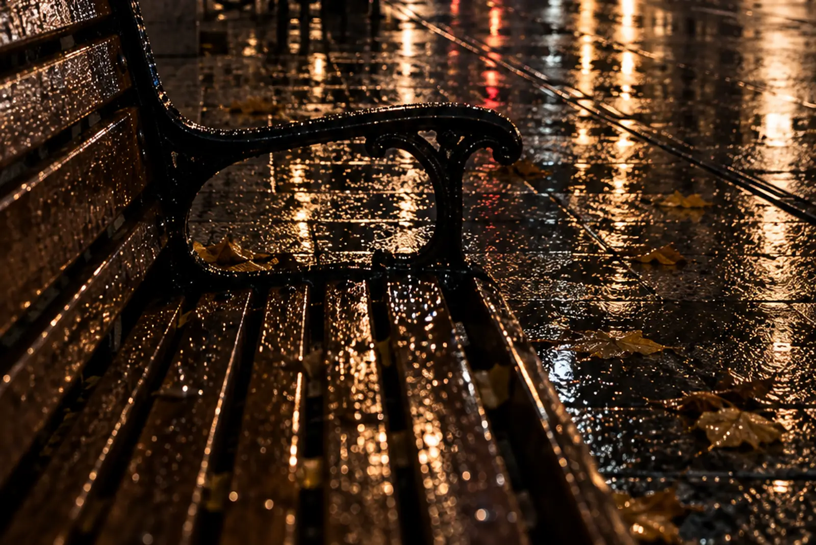 Eine leere Parkbank bei Nacht nach Regen, aus der Nähe: nasse Holzlatten und gusseiserne Armlehne im Laternenlicht, einzelne Herbstblätter darauf, rechts das nasse Pflaster.